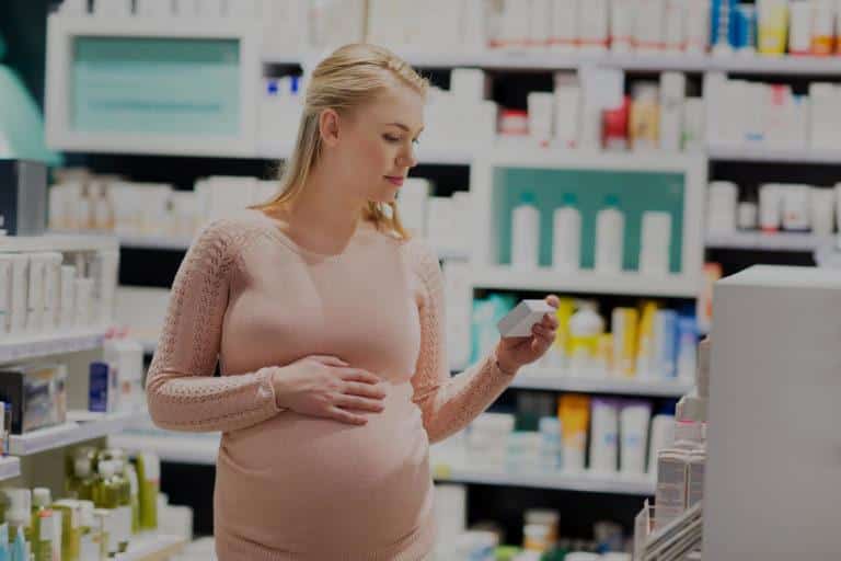 medicament grossesse risque Femme blonde enceinte qui marche dans une pharmacie et qui regarde la boite d'un médicament