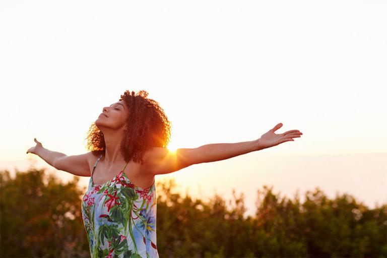 femme brune levant les bras devant un coucher de soleil